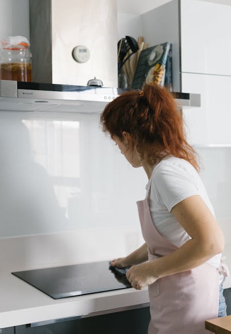 A woman with red hair, wearing a white t-shirt and a light pink apron, is engaged in surface cleaning on a black glass stove top in a modern kitchen. The kitchen features white cabinetry with a sleek, minimalist design, stainless steel appliances, and a white countertop. Visible in the background are a jar with cleaning supplies, a remote control, and kitchen utensils stored in a container. The area is well-lit with natural light, highlighting the cleanliness and shine of the surfaces, which appear free of dust and dirt. This scene exemplifies thorough domestic cleaning and surface sanitisation conducted by Oven Cleaning Ilford, emphasizing the importance of maintaining hygiene and cleanliness in a kitchen environment.