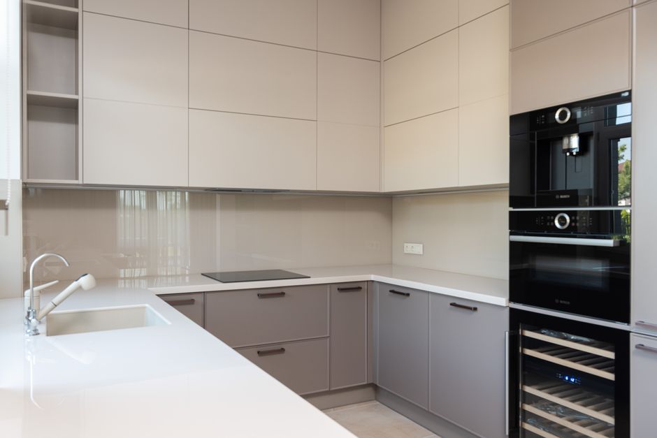A modern, L-shaped kitchen with smooth, light beige upper cabinets and darker gray lower cabinets, featuring minimalist handles. The kitchen surface includes a white quartz countertop with a built-in sink and chrome faucet, which appears clean and polished. The background shows a sleek, black induction cooktop embedded in the countertop and a wall-mounted electrical outlet. On the right side, there are two black, built-in appliances, likely an oven and microwave, integrated into the cabinetry. The room is well-lit with natural light, highlighting the shiny, tidy surfaces and the absence of debris or stains, demonstrating thorough surface cleaning and maintenance. This setup, along with the visible cleaning tools and appliances, emphasizes hygienic and deep cleaning standards typical of domestic surface cleaning services promoted by Oven Cleaning Ilford.
