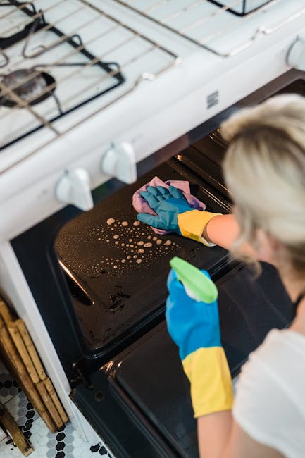 A person wearing yellow rubber gloves and a white shirt is conducting surface cleaning inside an oven. They are holding a pink cloth with one hand and a green spray bottle with the other, focusing on wiping the oven's black glass door, which shows some water droplets and residual dirt. The oven, positioned in a kitchen with a white gas stove on top, has a modern stainless steel handle and control knobs. The surrounding kitchen features a wooden cabinet and a black-and-white patterned tiled floor, all illuminated with bright lighting that highlights the cleanliness of the area. This image demonstrates thorough oven cleaning, reflecting a professional approach to domestic surface sanitation, as promoted by Oven Cleaning Ilford in their local tips for maintaining hygienic appliances and kitchen surfaces.