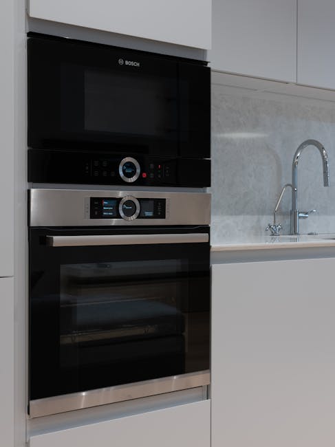 A modern, L-shaped kitchen with smooth, light beige upper cabinets and darker gray lower cabinets, featuring minimalist handles. The kitchen surface includes a white quartz countertop with a built-in sink and chrome faucet, which appears clean and polished. The background shows a sleek, black induction cooktop embedded in the countertop and a wall-mounted electrical outlet. On the right side, there are two black, built-in appliances, likely an oven and microwave, integrated into the cabinetry. The room is well-lit with natural light, highlighting the shiny, tidy surfaces and the absence of debris or stains, demonstrating thorough surface cleaning and maintenance. This setup, along with the visible cleaning tools and appliances, emphasizes hygienic and deep cleaning standards typical of domestic surface cleaning services promoted by Oven Cleaning Ilford.