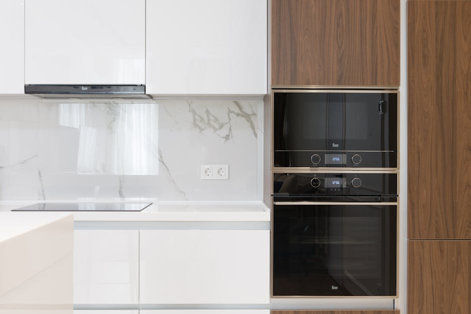 A modern kitchen with white cabinetry and a gray countertop, featuring a built-in stainless steel oven with a glass door. Above the oven, there is a stainless steel range hood mounted on the wall. To 