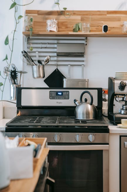 A modern kitchen scene featuring a stainless steel oven and stovetop with a kettle on one of the burners, situated beneath a wooden shelf. The shelf holds glass jars, a small potted plant, and other items, while a metal railing with hooks above the stove displays utensils and a black oven mitt. The white countertop and backsplash have a clean appearance, with minimal clutter, emphasizing surface cleaning and maintenance. The bright, natural lighting highlights the shiny, reflective surfaces of the oven and stainless steel appliances, conveying a sense of hygiene and deep cleaning. This setting exemplifies domestic cleaning efforts by Oven Cleaning Ilford focused on surface sanitisation and routine upkeep of kitchen appliances and surfaces.