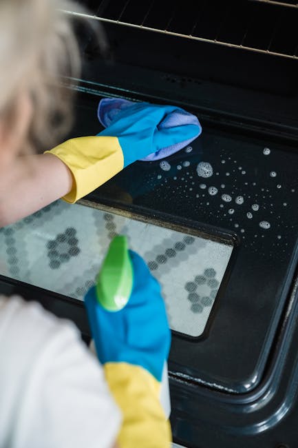 Close-up of a person wearing yellow and blue rubber gloves cleaning the interior of a black oven with a wire rack, which is being pulled out to reveal a clean, shiny ceramic and stainless steel surfac