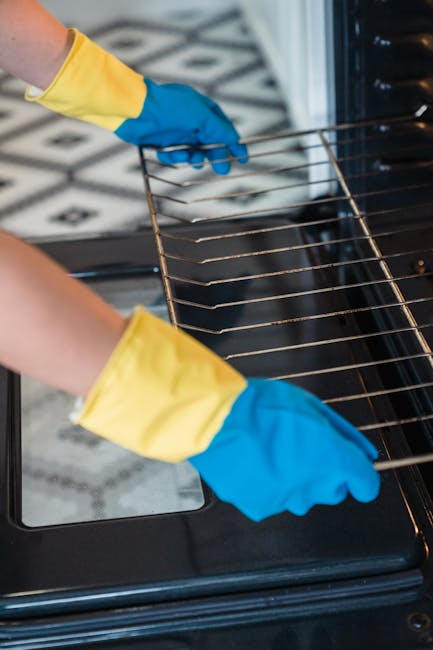 Close-up of a person wearing yellow and blue rubber gloves performing surface cleaning inside a black oven. The individual is holding a metal oven rack over the oven's interior, which appears clean and free of grease or debris. The oven's interior is illuminated, showing shiny, reflective surfaces with no visible residue. The background features a patterned tiled floor, and the scene emphasizes the meticulous care involved in deep cleaning and sanitisation, as offered by Oven Cleaning Ilford, the company specializing in domestic cleaning services tailored for busy landlords on Ilford High Road.