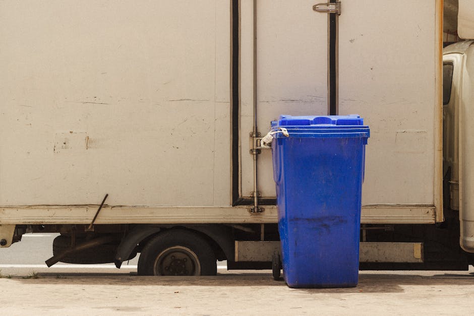 An overflowing public collection point with multiple waste bins and numerous bags and cardboard boxes scattered on the pavement, containing mixed paper, cardboard, plastic, and general waste; behind t